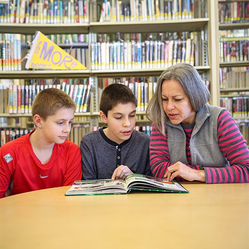 a parent and two school-aged children sit in the library with a book in front of them