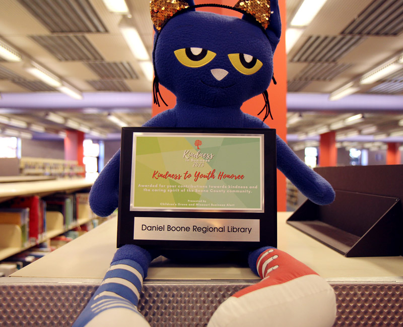 Kindness to Youth Award plaque for the Daniel Boone Regional Library sits in the lap of a large Pete the Cat stuffed animal on top of library shelves