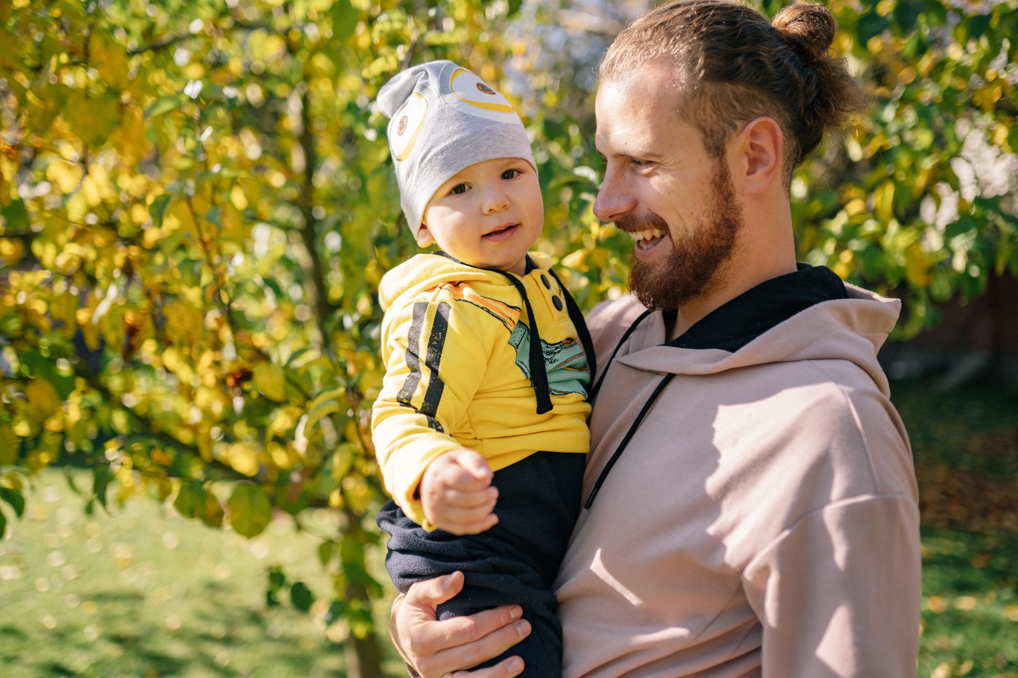 A smiling father holds his toddler as they look at fall leaves. 