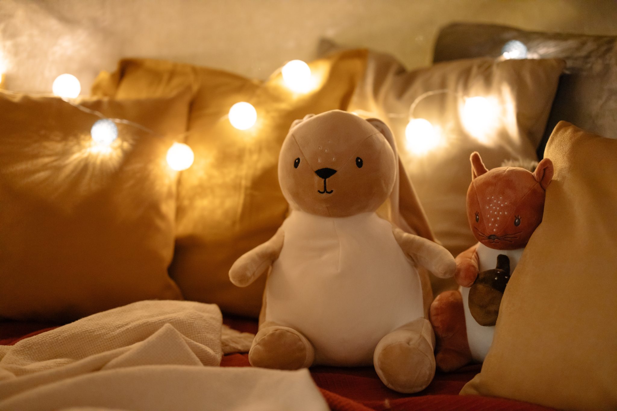 A stuffed bear and a stuffed squirrel sit in a cozy pile of blankets and pillows. Round string lights are draped across the scene. 