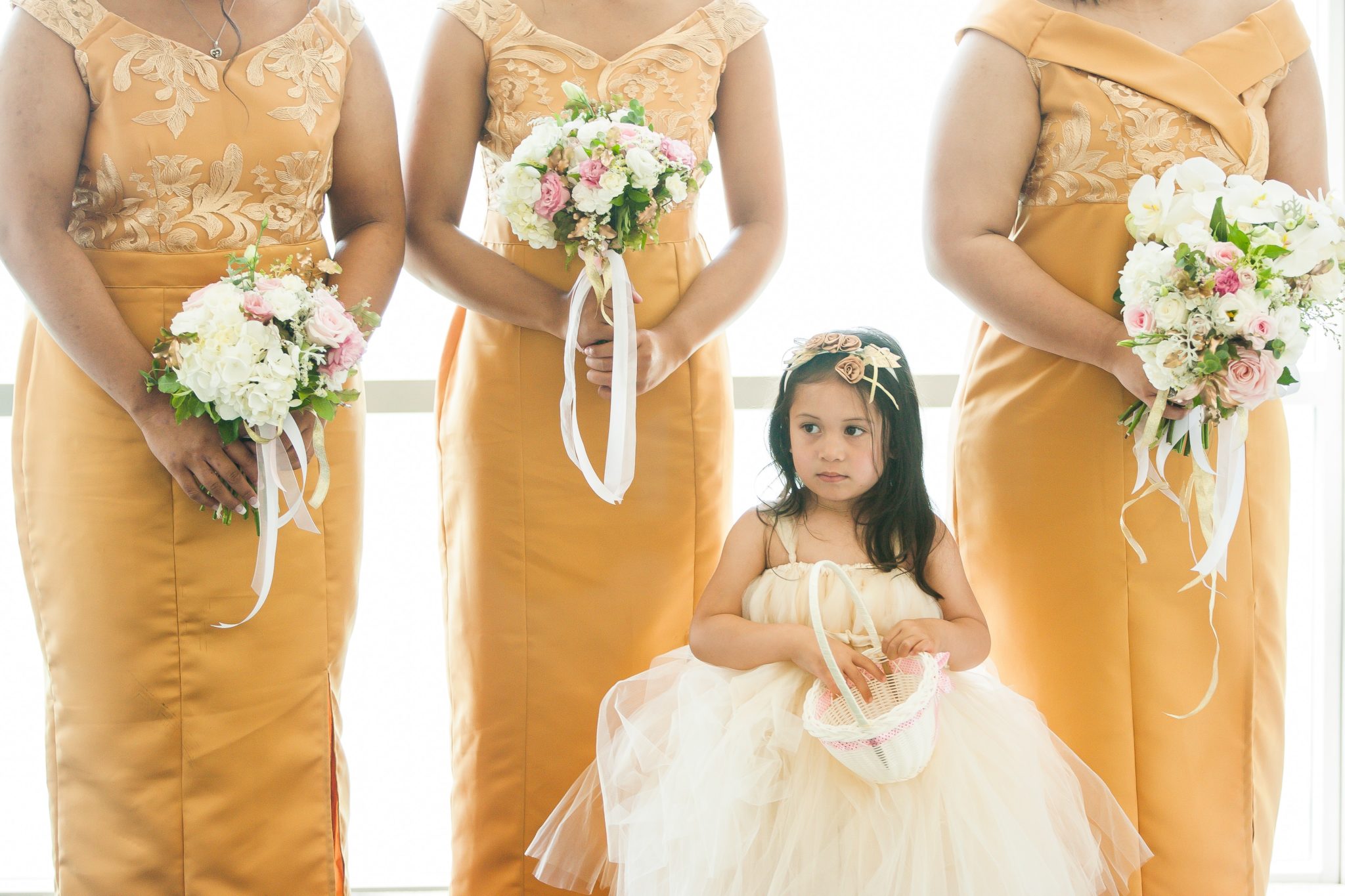 A young flower girl in a white dress stands in front of a line of bridesmaids in yellow dresses. The flower girl holds a basket of petals. 