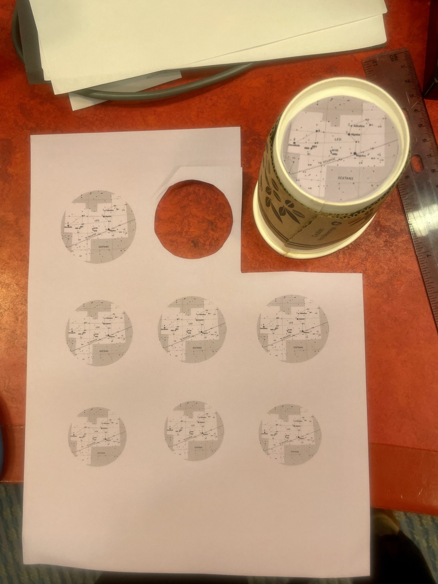 Image of purple/gray sheet of paper with star-maps printed on it, situated next to a paper coffee cup turned upside down. The top right corner of the sheet is cut off and a circle is cut out of the top center. A circular map (cut out from the top-center of the sheet) is placed on the bottom of the upside-down cup to demonstrate the correct sizing needed for the map if using a paper cup for this craft.