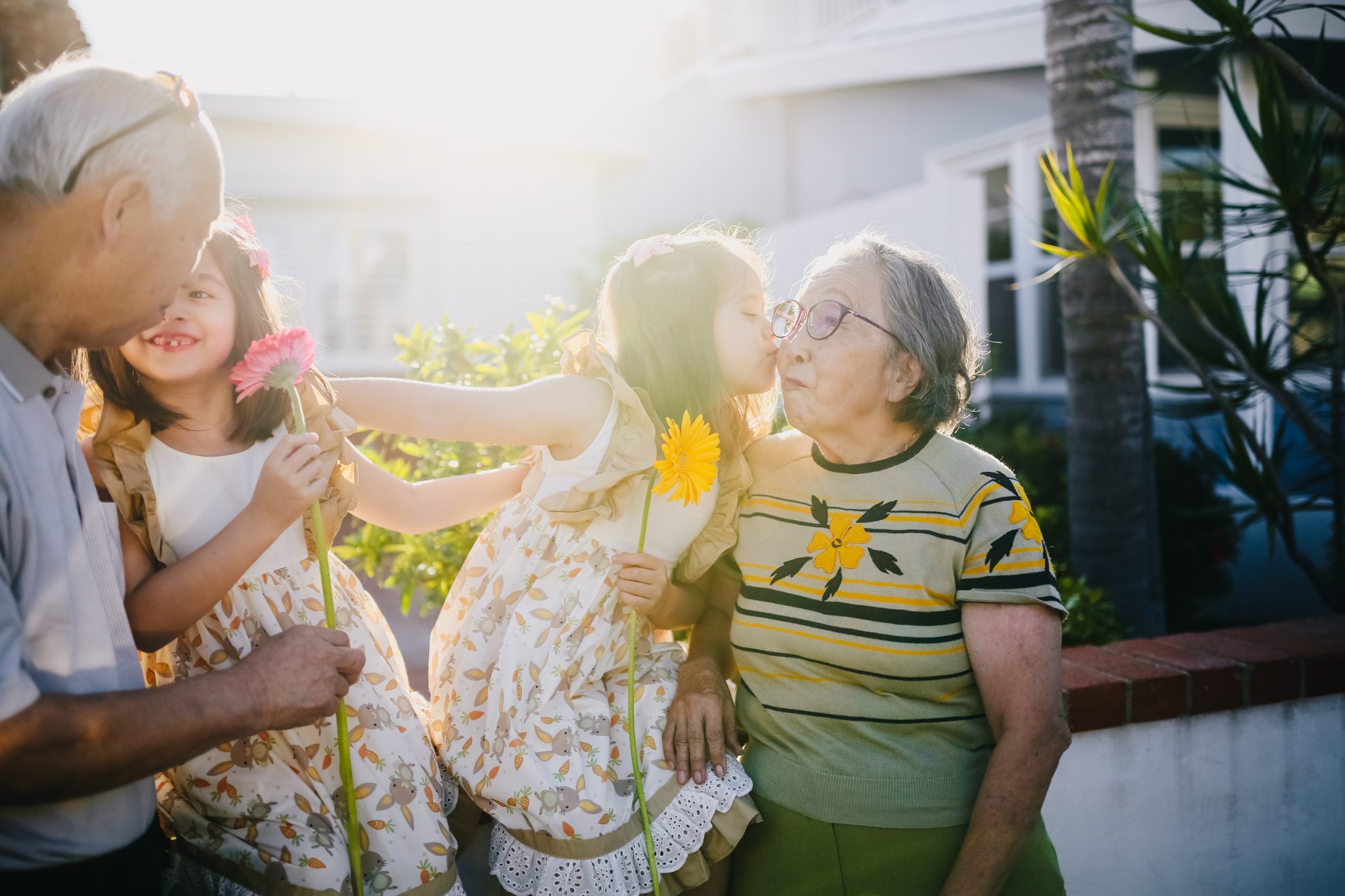 Two young children in sundresses sit on a low wall outdoors while holding flowers. An elderly man and woman stand beside them. The children offer kisses to their grandparents. 