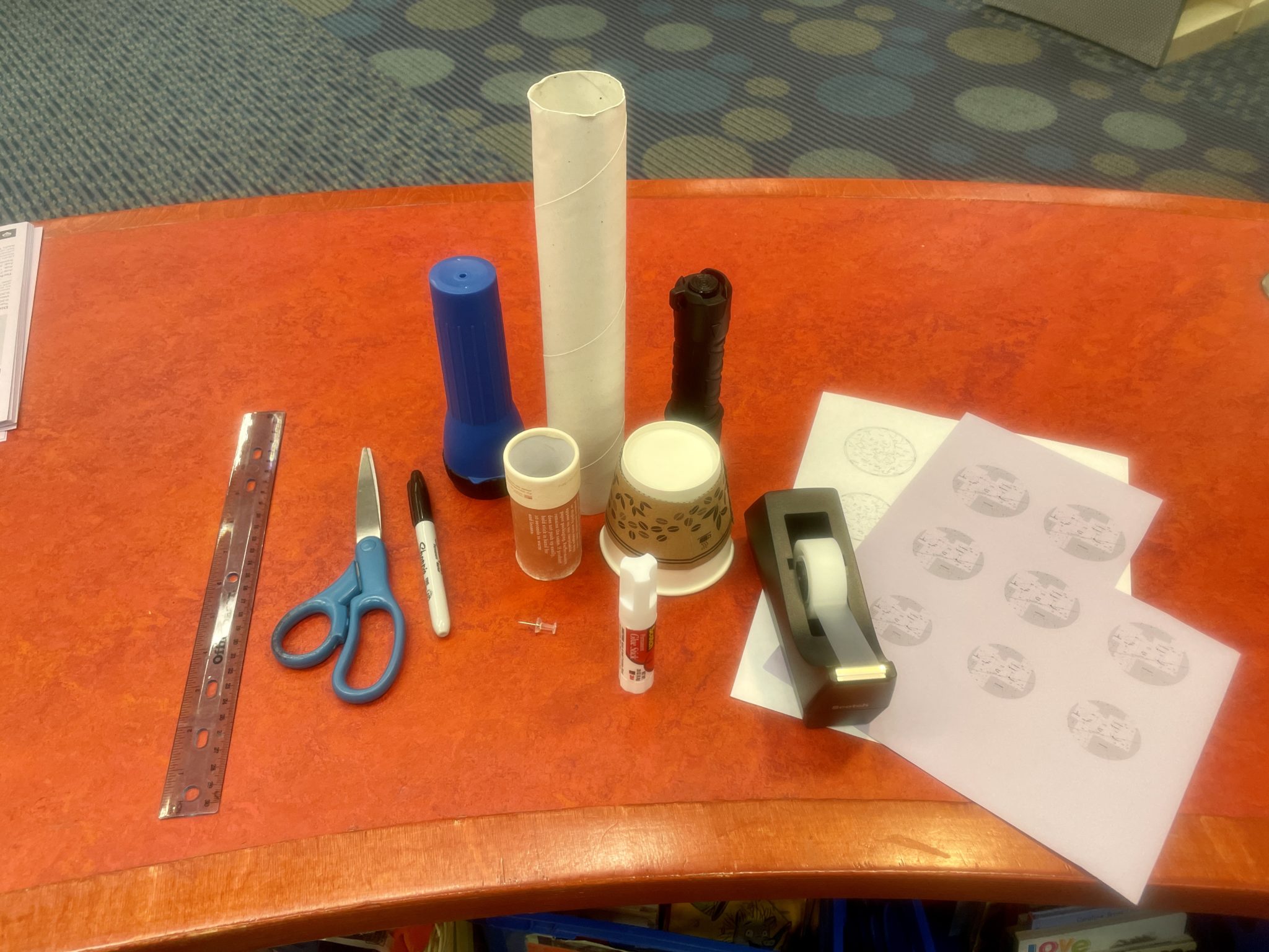 Image of materials needed to make this craft, including a ruler, pair of scissors, sharpie, push pin, 2 flashlights (one blue, one black), 2 cardboard tubes (one tall, one short), a paper coffee cup, tape/tape dispenser, and two printed out sheets featuring circular star-maps.