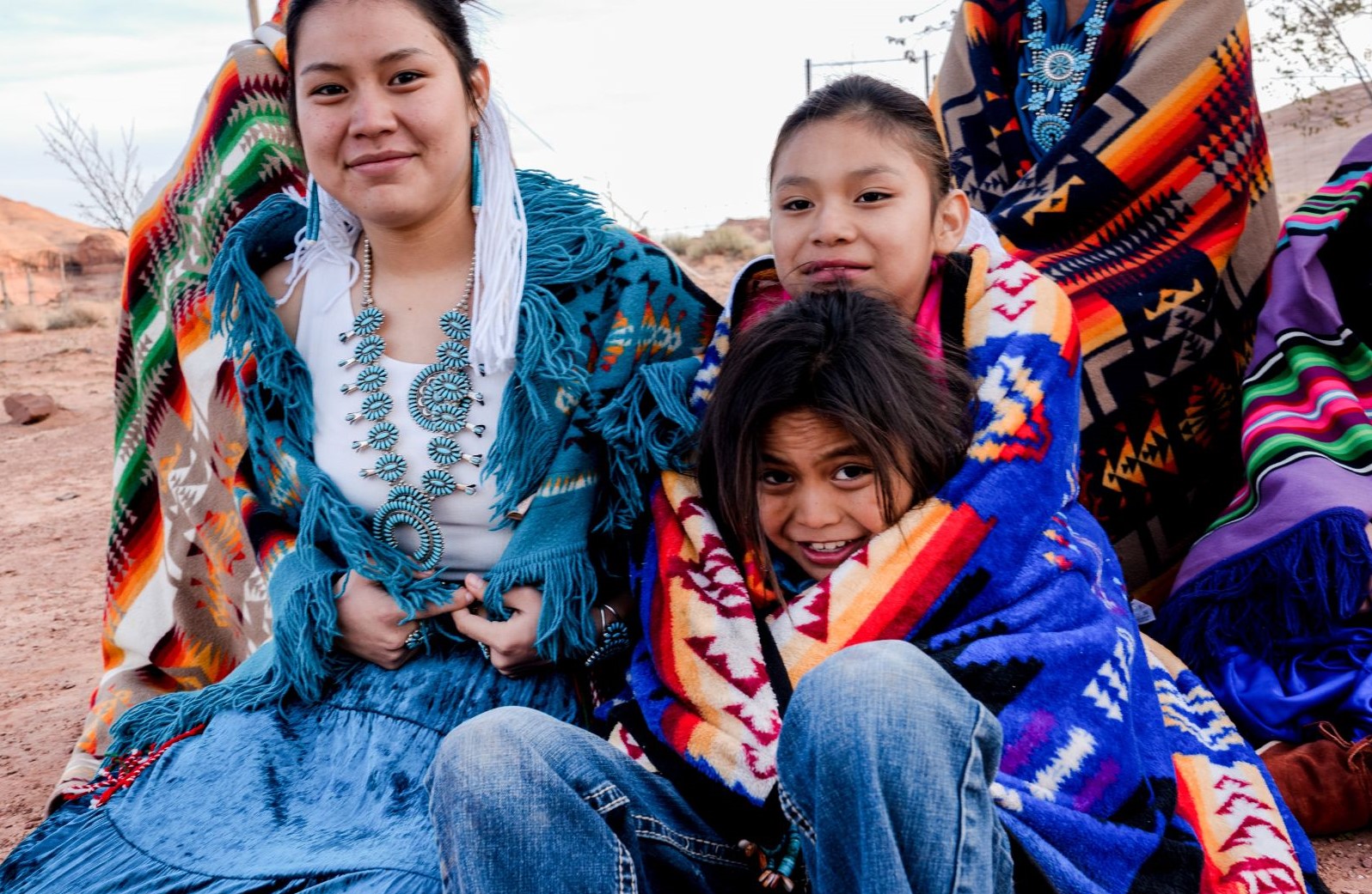 Three Navajo young people sit outside on the ground. A teen sits beside two children with their arms wrapped around each other. All three are wearing Navajo blankets and jewelry. 