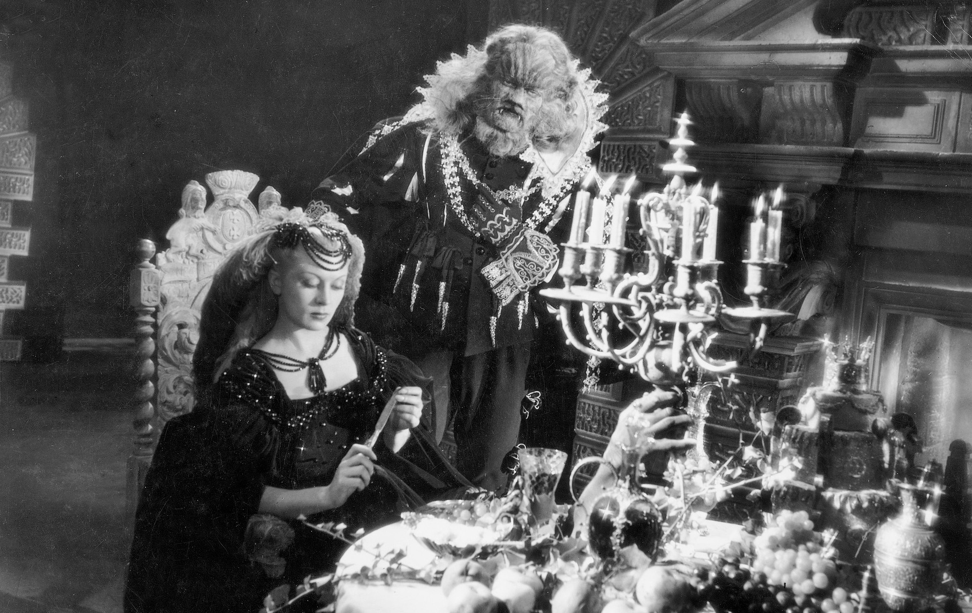 Film still featuring Beauty (Belle) fiddling with a butter knife while seated at a table piled high with various food, drink and ornate tableware. The Beast stands ominously behind her.