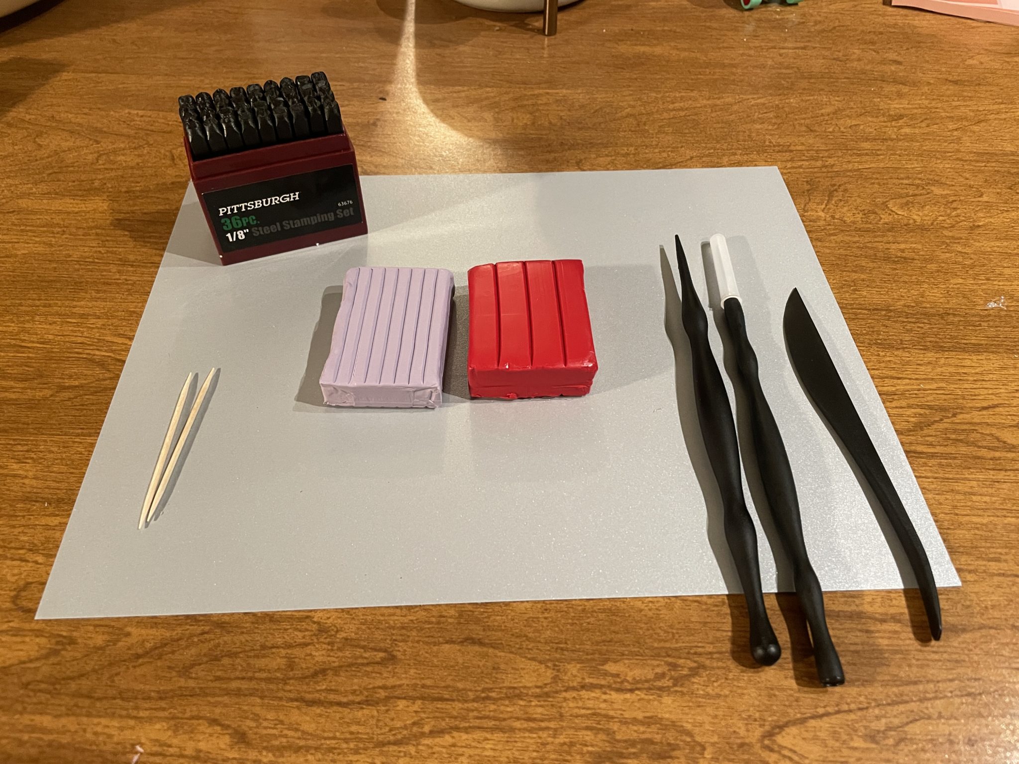 Photo of materials needed to make your own clay conversation heart: a silver sheet of construction paper (i.e., clean work surface), two toothpicks, a set of metal alphabet stamps, two blocks of oven-bake polymer clay (one in lilac, the other in red), and three black plastic clay sculpting tools including a plastic craft knife.