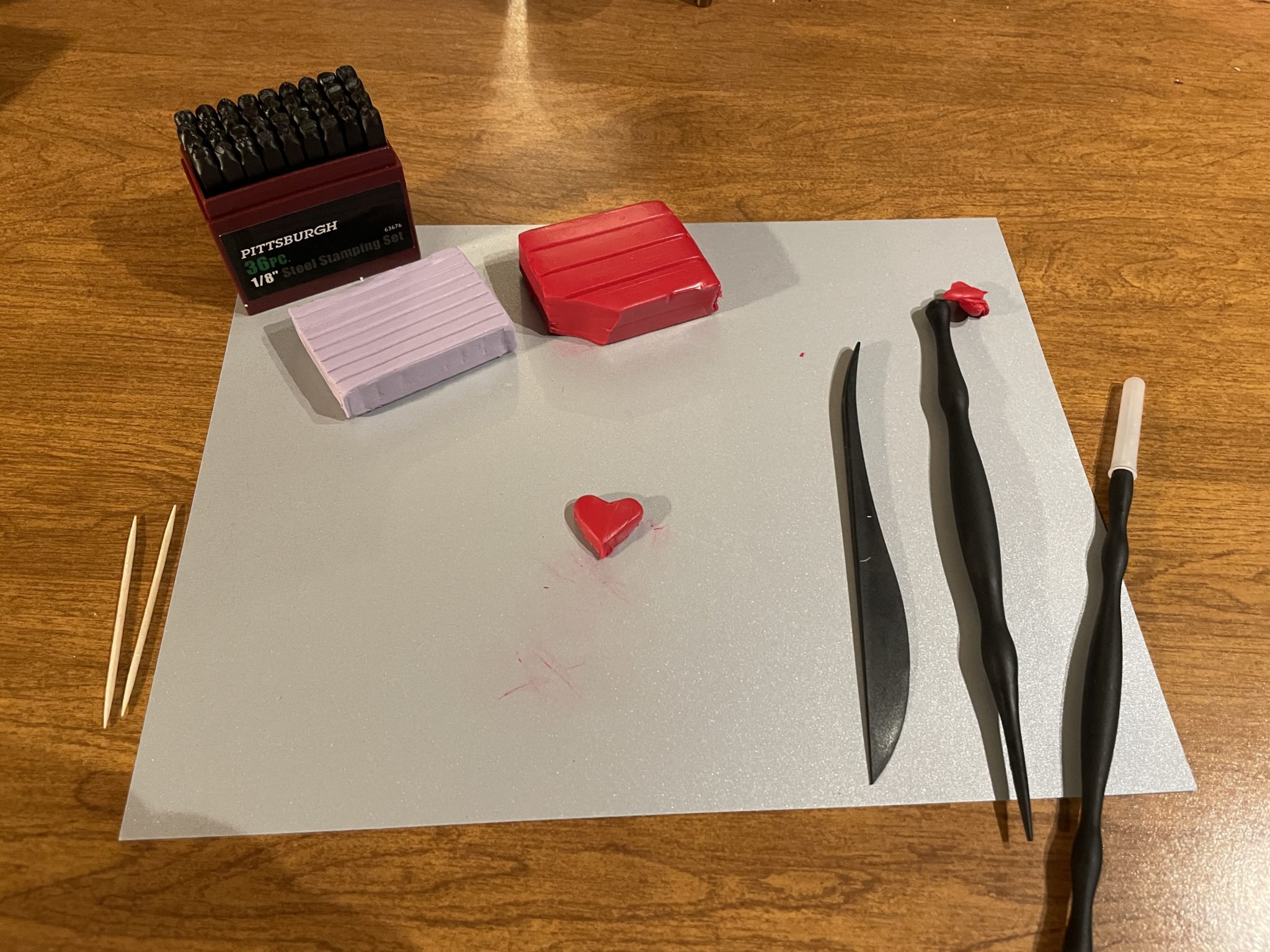 Photo of materials needed to make your own clay conversation heart. The small red clay ball has been shaped into a heart and placed in the center of the silver construction paper. 
