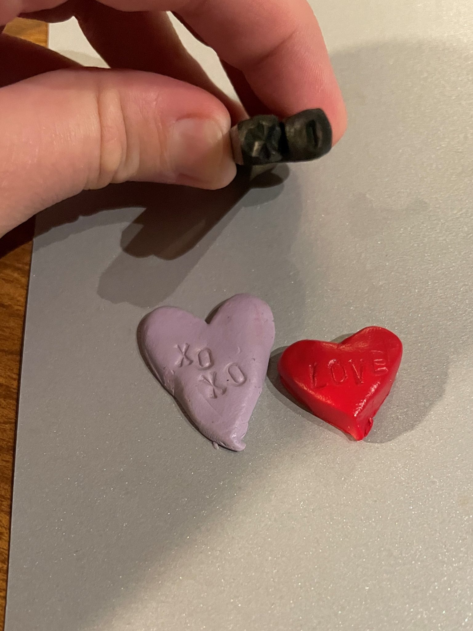 Close-up photo of two metal alphabet stamps (the letters "X" and "O") held above two clay hearts: the bigger lilac heart has the phrase "XOXO" stamped on it while the smaller red clay heart has the word "LOVE" stamped on it. 
