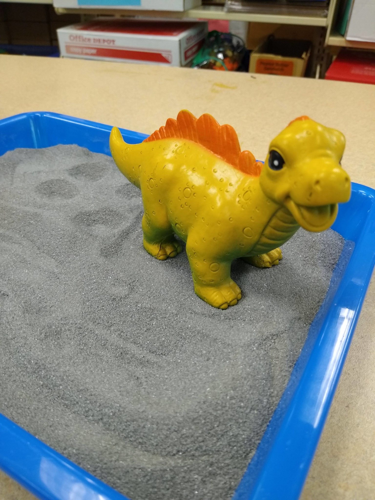 A smiling yellow dinosaur toy standing in the sand tray. Footprints and a stream have been drawn in the sand. 