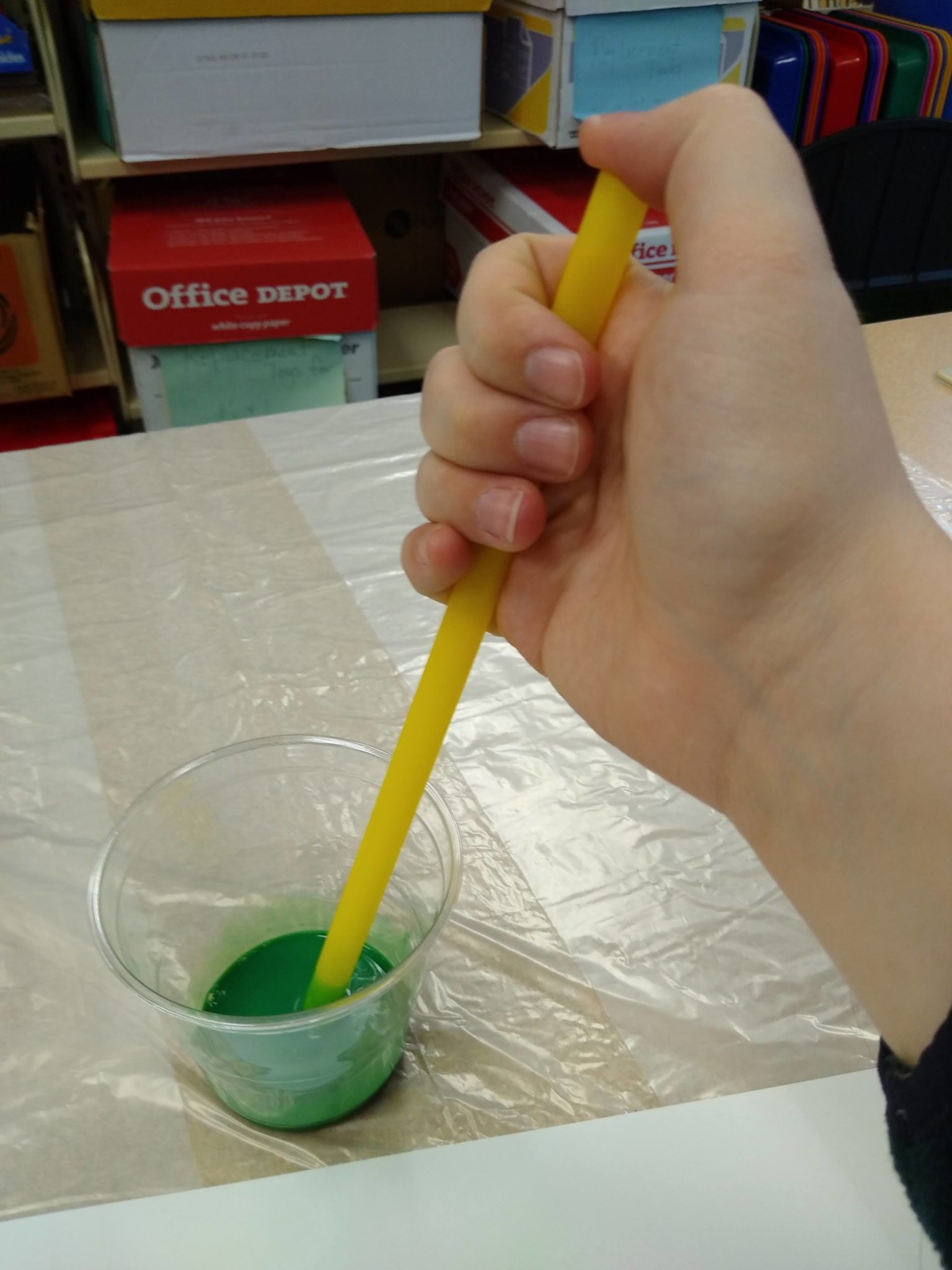 Placing a straw in a small cup of green paint and placing a thumb over the top of the straw.