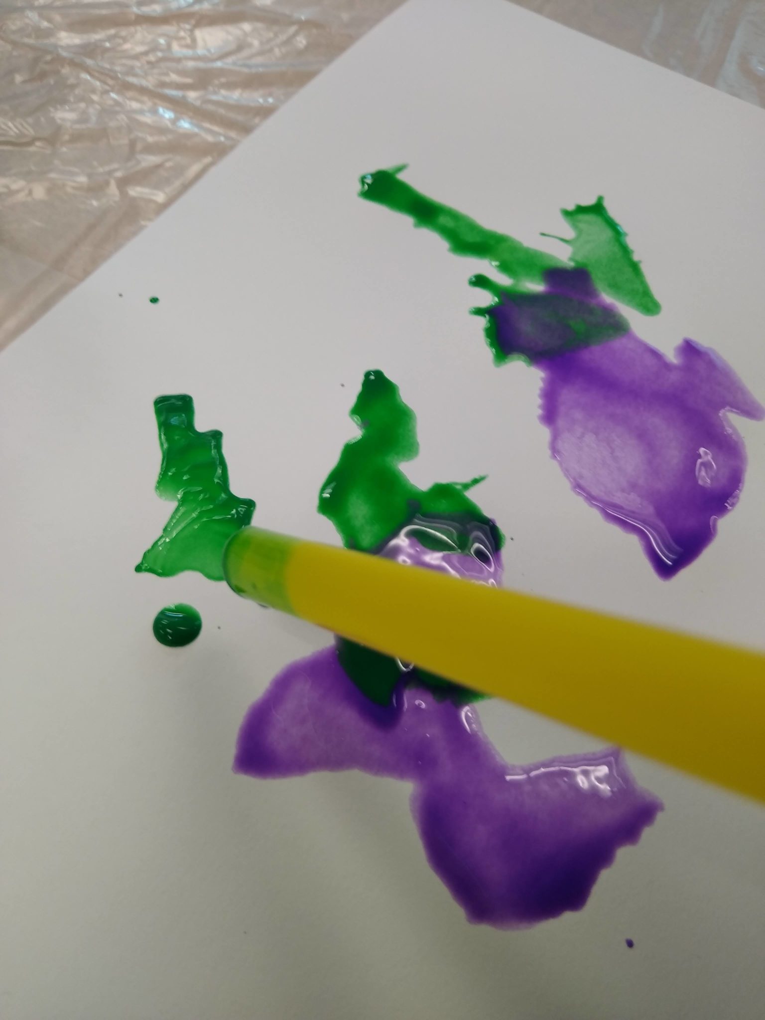 Looking down at a straw pointed at purple and green paint splotches on paper.