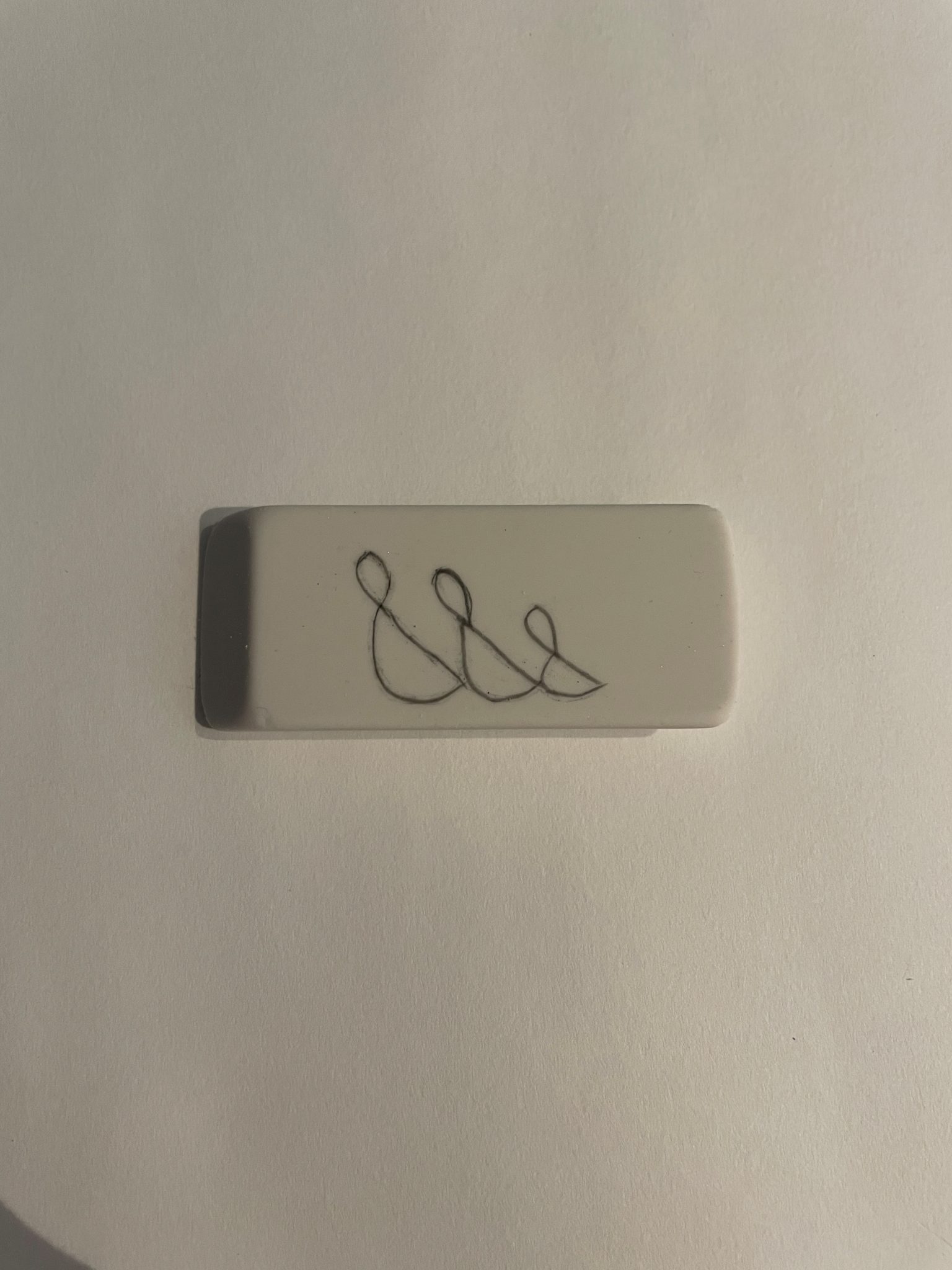 Close-up of a white rectangular eraser with an abstracted design marked in the center of the eraser with pencil. The abstracted design looks like three connected geese bodies and heads arranged by size with the largest goose body/head appearing on the left and the smallest goose body/head on the right. 