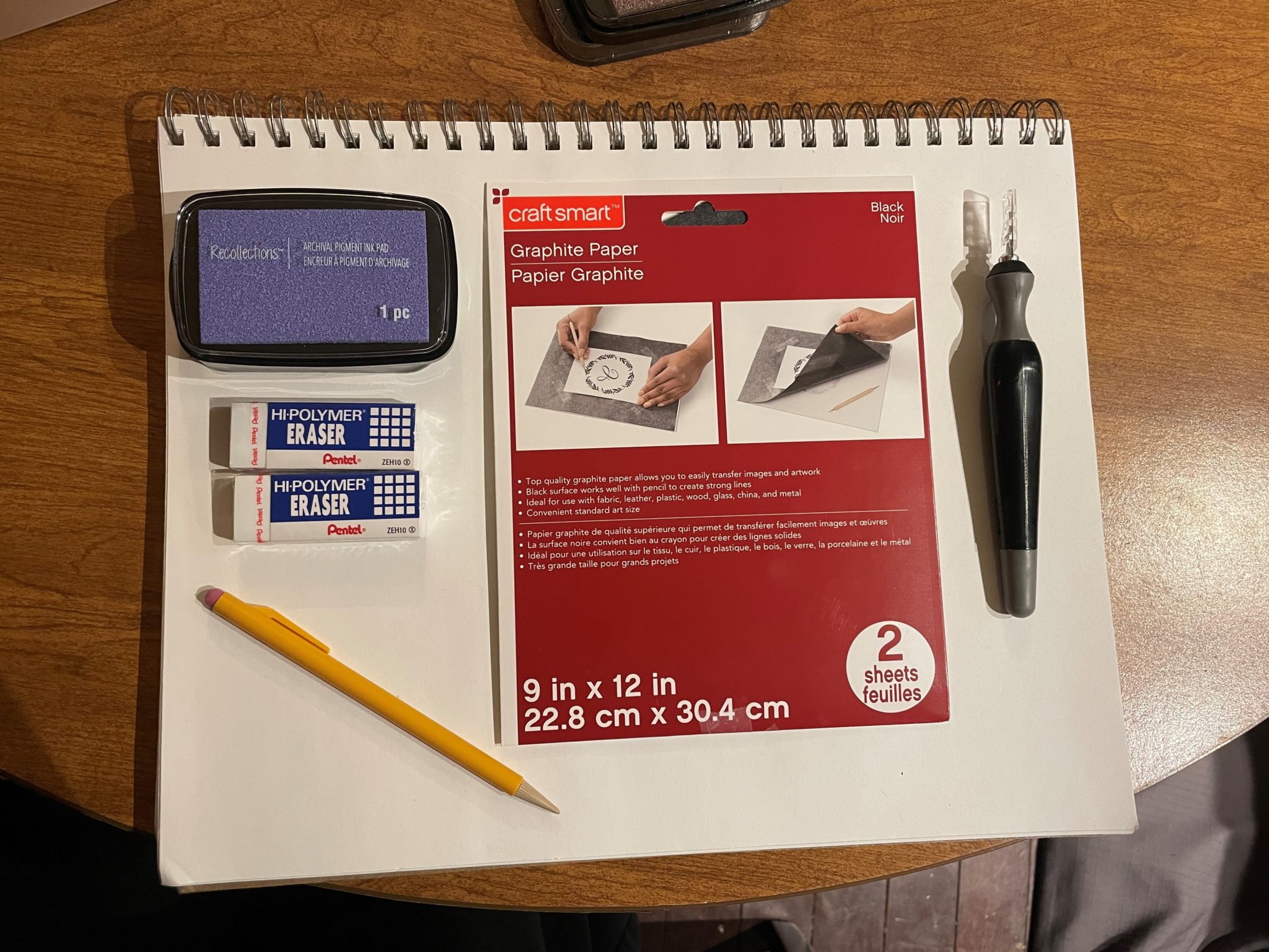 Materials for making an eraser stamp are arranged on a white sketchbook page: a lilac ink pad, two white rectangular erasers, a mechanical pencil, a sheet of graphite paper, and an x-acto knife.