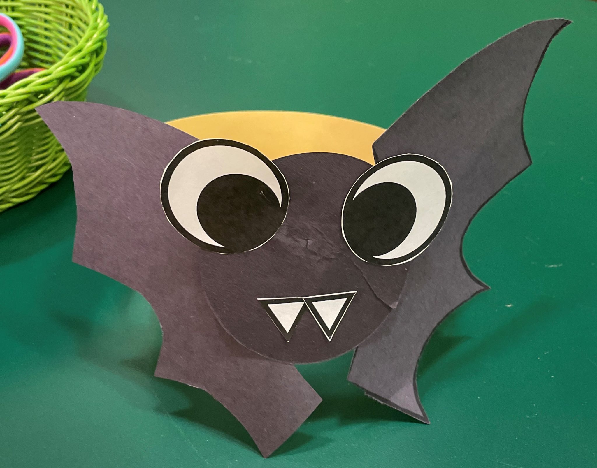 Photo of a papercraft bat headband on a green desk. The paper bat's face and wings are made of black construction paper with white fangs and huge black and white googly eyes. The bat face and wings are attached to a yellow strip of paper glued in a circle. The eyes and wings are attached a little askew and there are no ears, creating an adorable misshapen look.