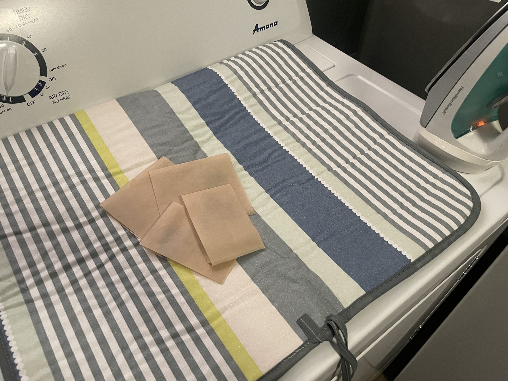 Photo of a striped ironing blanket with four pieces of folded waxed paper laid on top. The ironing blanket is laid on top of a dryer next to an iron.