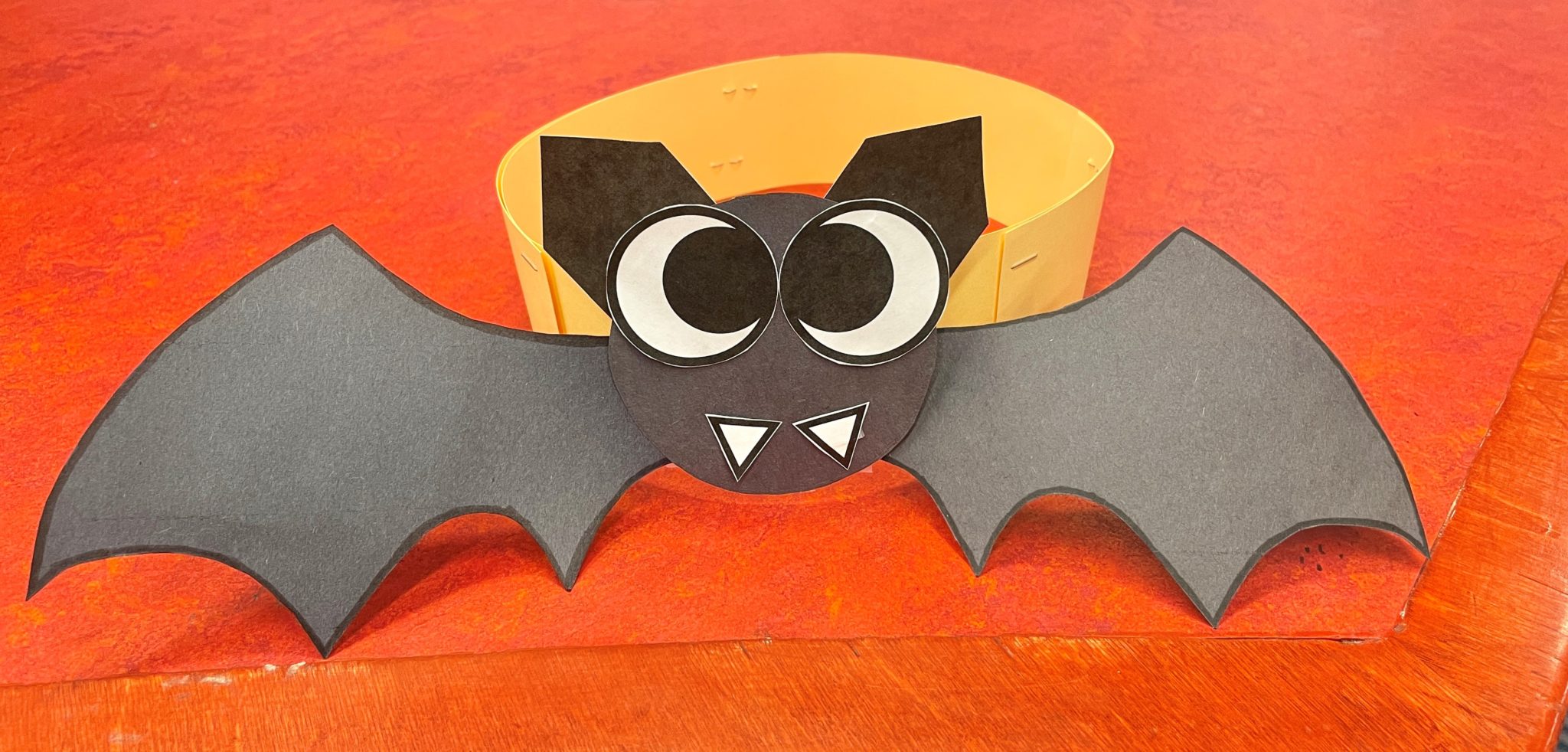Photo of a papercraft bat headband on a red desk. The paper bat's face, ears, and wings are made of black construction paper with white fangs and huge black and white googly eyes. The bat face and wings are attached to a yellow strip of paper stapled in a circle.