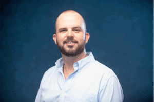 headshot of a man wearing a light blue shirt in front of a blue background