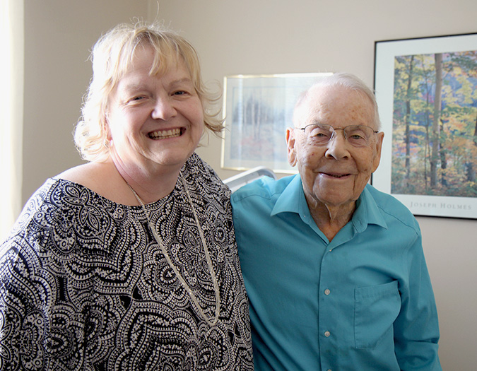 Carolyn and Cletus stand closely and smile in his apartment facing the camera