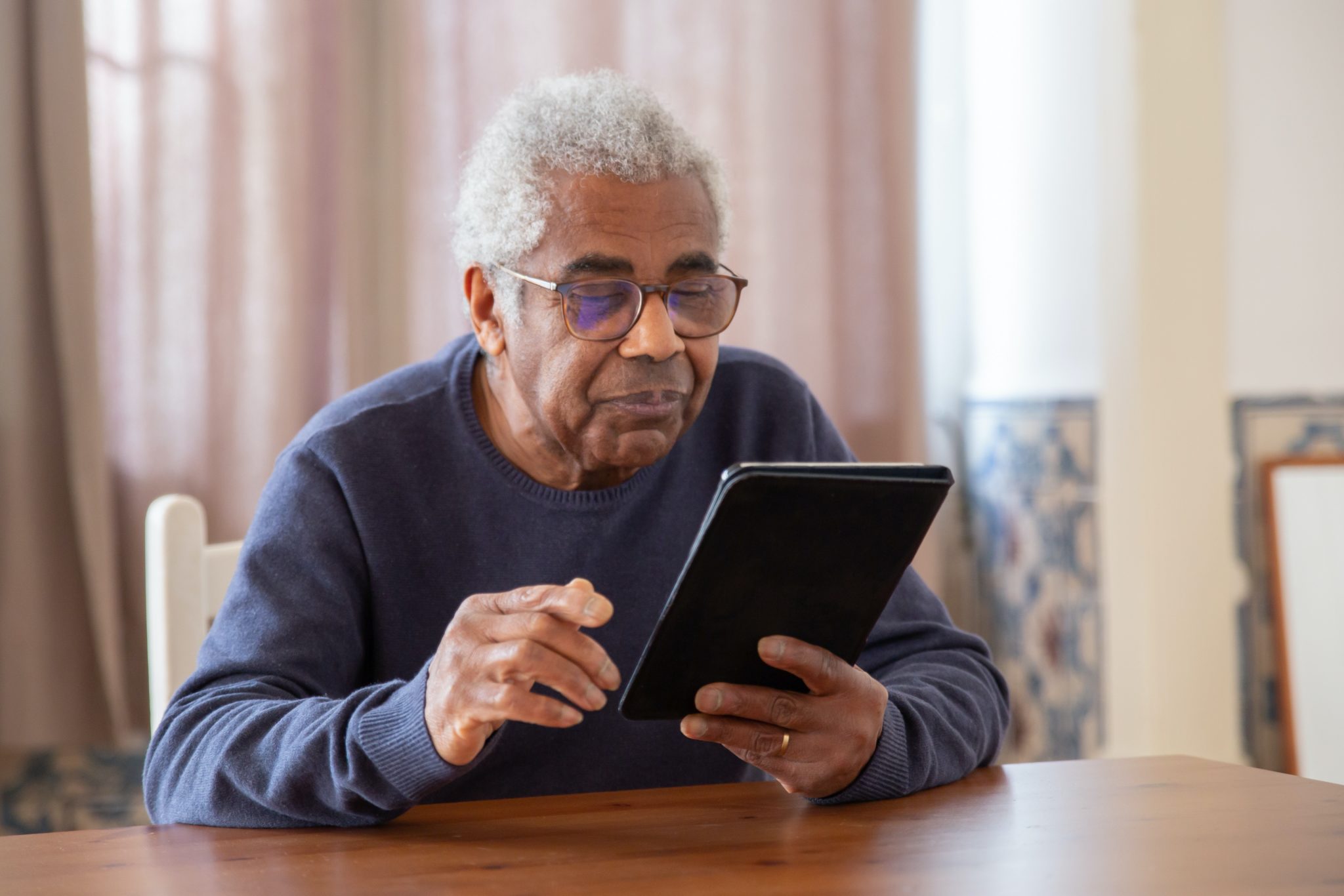 An elderly Black man in a blue sweater using a digital tablet
