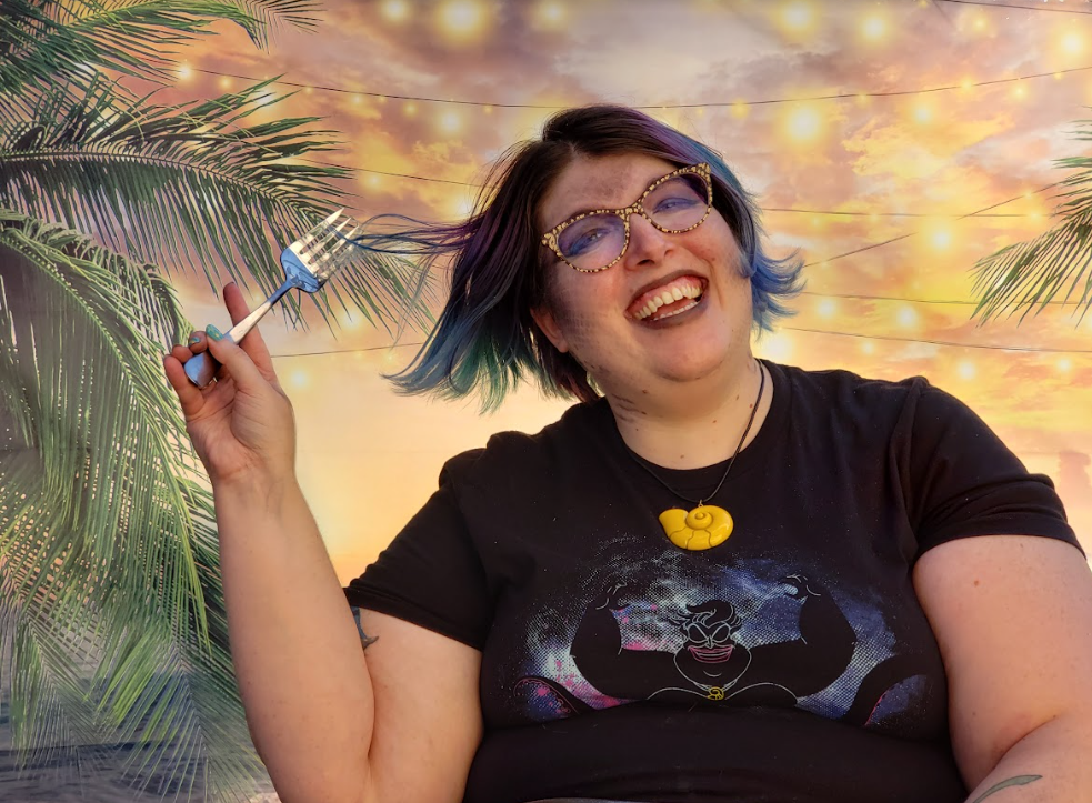 Woman with short blue hair and glasses with painted on scales combing her hair with a fork and wearing an Ursula shirt. 