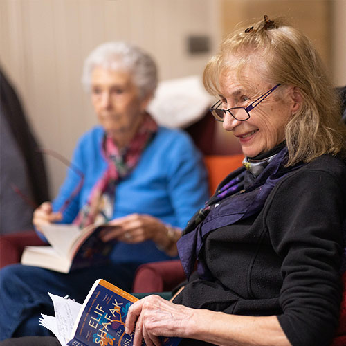 A woman in the foreground holds an open book and a pencil, smiling and looking towards other members of a book discussion group