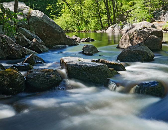 a small river runs over rocks with green trees in the background reflected on smooth water