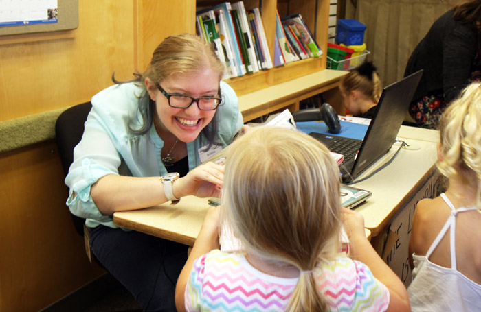 On board Bookmobile, Jr., a library staff member leans across a small desk to talk to a small child checking out books