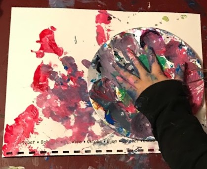 Child's hand inside a messy paint palette, on top of calendar page covered in paint. 