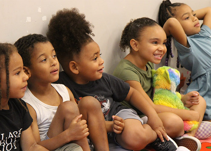 preschool children sit with their backs against the wall, smiling off camera