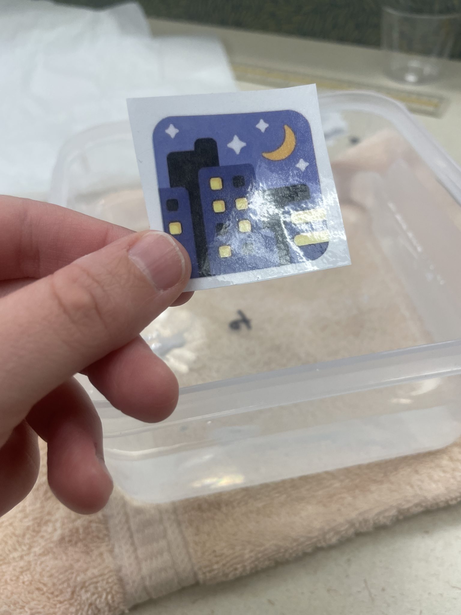 Image of a white hand holding a toner-printed image of the night with stars emoji that has been covered in a layer of packing tape, making the image a little glossy. The hand holds the taped-up image over a small tub of water, preparing to drop it in. 