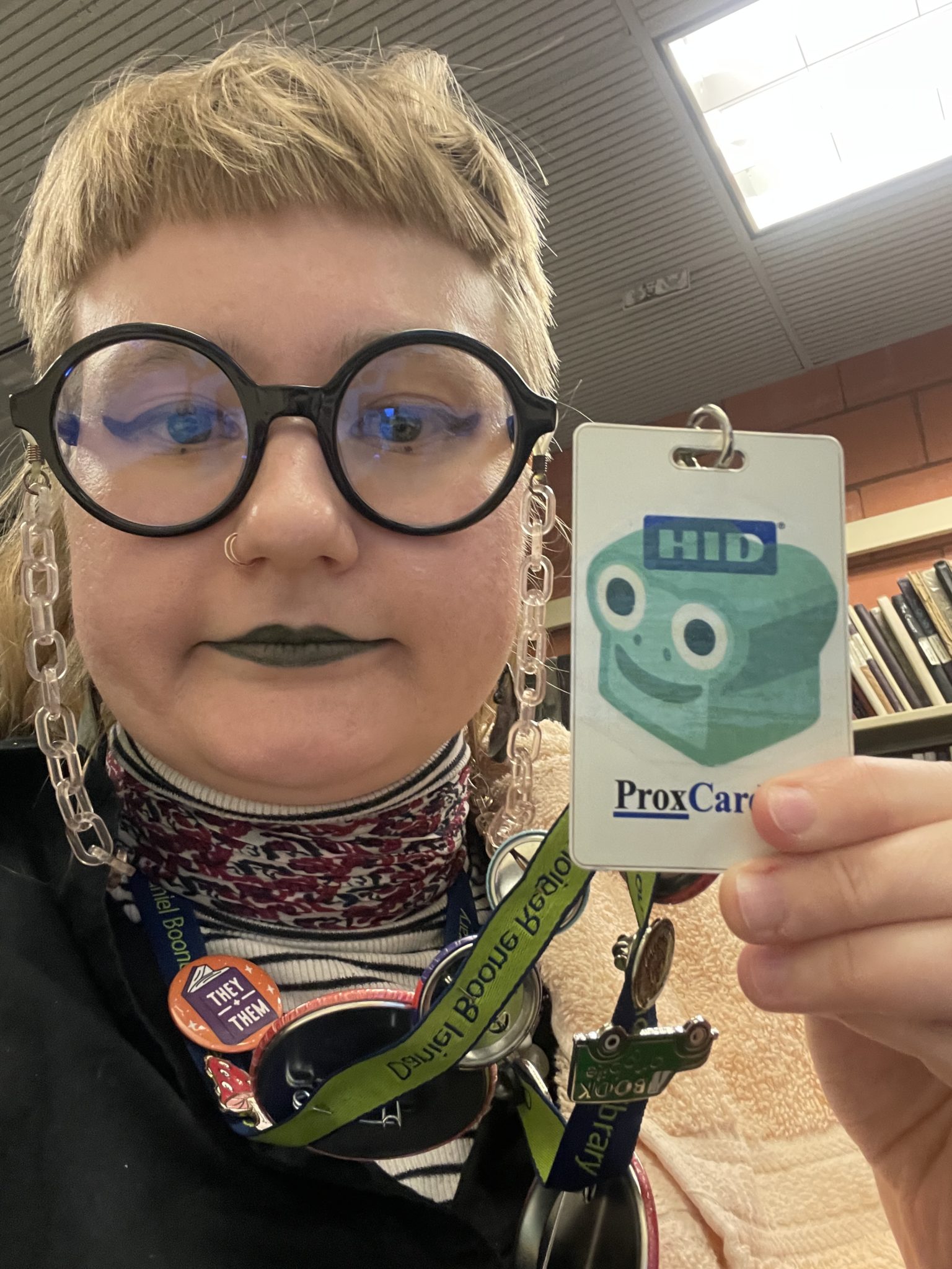 Image of Grae, a white nonbinary person, holding up a library staff keycard to which a packing tape sticker is affixed. The packing tape sticker on their keycard is an emoji fusion of the frog and bread emojis, creating a cute green loaf of bread with a wide-eyed frog's face superimposed on the front of the loaf. 