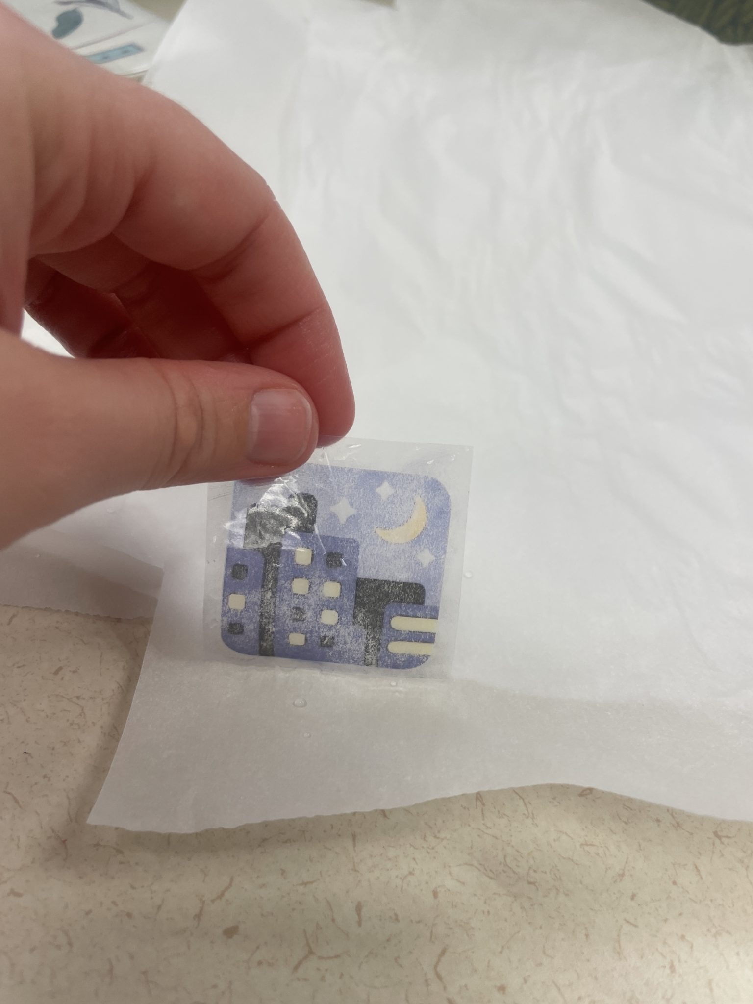 Image of a white hand laying a piece of wet packing tape dyed with toner -- aka a DIY sticker -- onto a sheet of wrinkled non-porous parchment paper. The toner on the packing tape shows the night with stars emoji. 