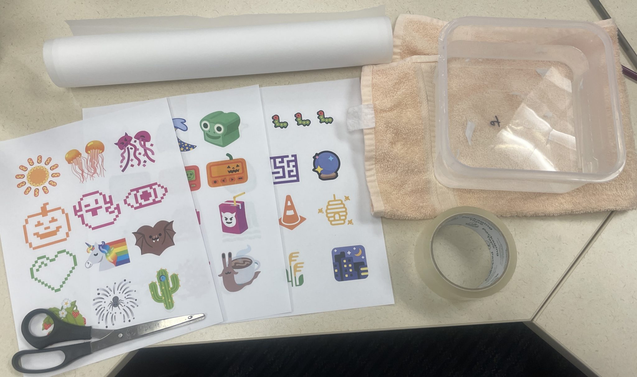Photograph of the materials needed to make this craft including clockwise from top: two sheets of non-porous parchment, a tea towel, a plastic tub of warmish water, a roll of clear packing tape, three pages of toner-printed images and a pair of scissors. 
