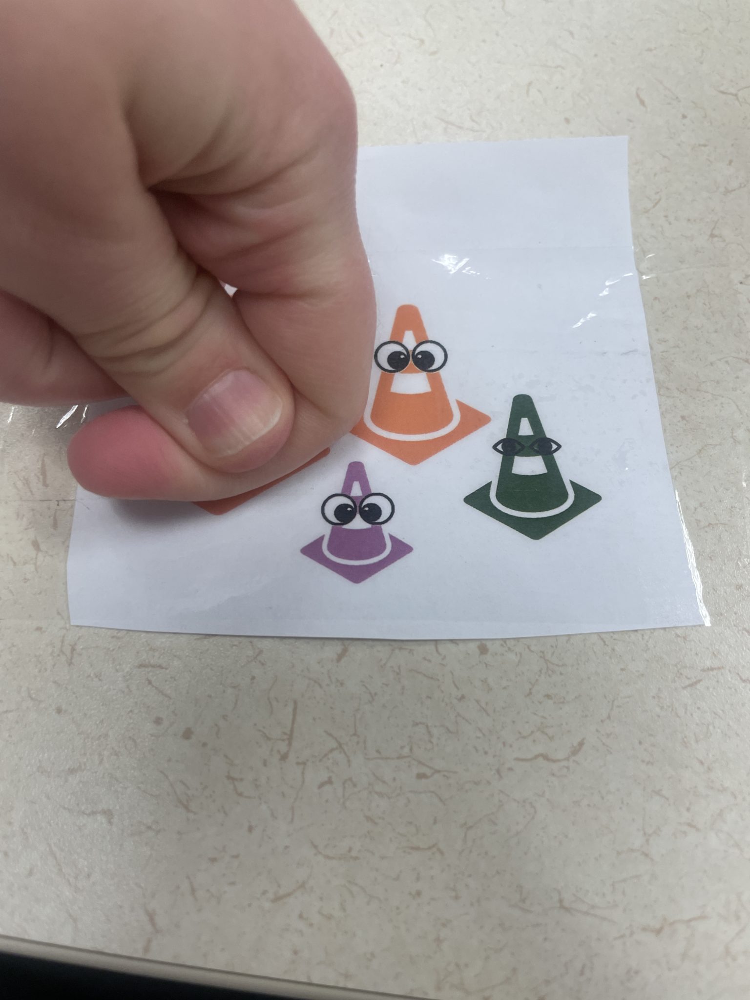 Image of a white hand pressing down on the first two knuckles of its pointer finger to burnish or rub packing tape across the top of a toner-printed image of four traffic cones with googly eyes (2 orange cones, 1 purple cone and 1 green cone).