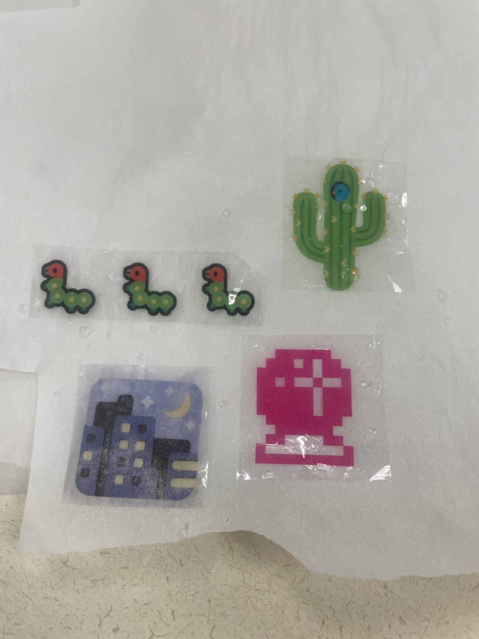Image of four wet packing tape stickers drying on a piece of wrinkled non-porous parchment paper. Clockwise from top right: a sticker of a blue bird poking out from a hole in a cactus; a sticker of a hot pink pixelated crystal ball; a sticker of the night with stars emoji; a sticker of three bug emojis printed in a line. 
