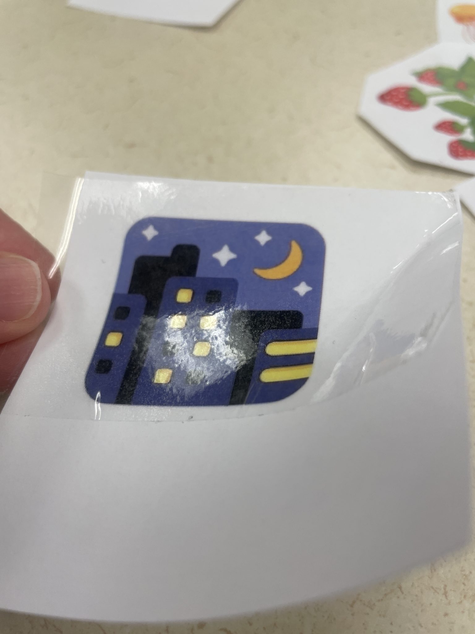 Photograph of a white hand carefully laying a piece of packing tape over a toner-printed image of the night with stars emoji, making sure there are no creases or air bubbles in the tape as it is laid over the image. 