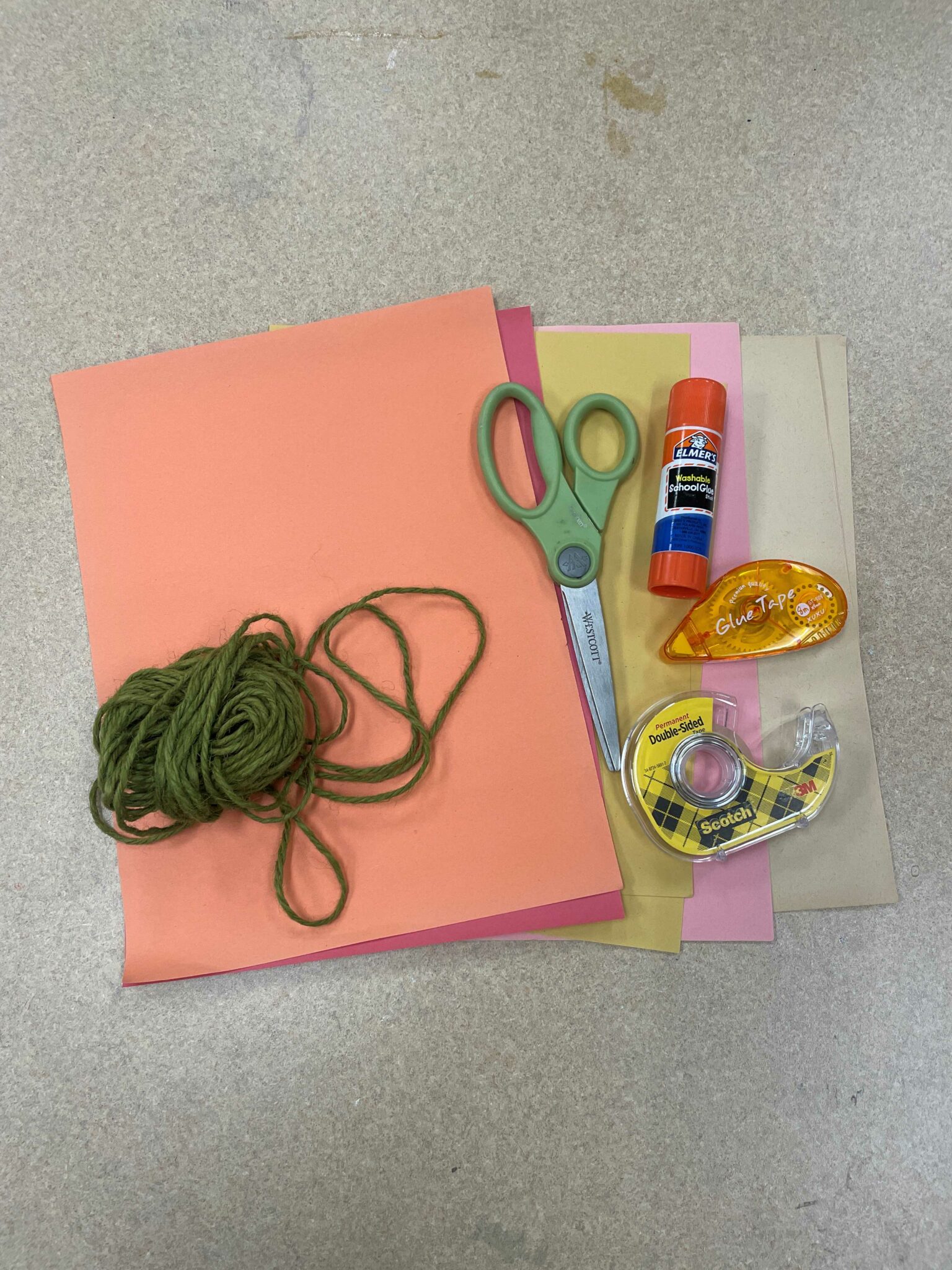 Photo of various art supplies for the craft outlined in this blog including a stack of colored paper (in orange, red, yellow, pink and brown), a bundle of green twine, a pair of green scissors, and various adhesives, including a glue stick, a roll of double-sided tape, and a glue tape dispenser. 