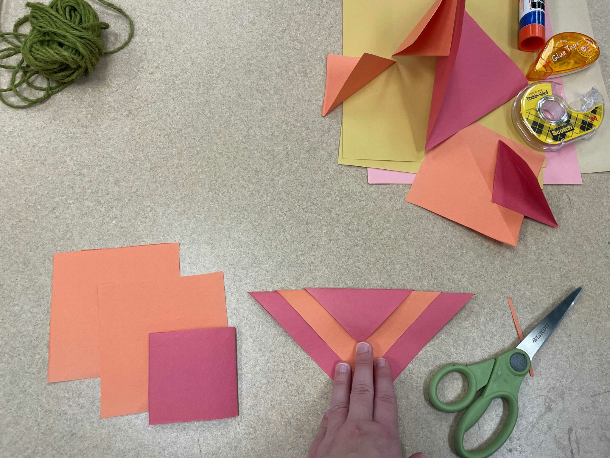 A white hand holds down three paper triangles layered on top of one another to create a red-orange-red colored flag. The triangles are arranged in a downward-pointing chevron pattern. In the background are two orange and one red squares of paper, which, when folded in half, could create the same triangle flag shape. There are other orange and red squares folded into triangles in the background as well, along with a ball of green twine, a pair of green scissors, and a variety of adhesives, including a glue stick, roll of double-sided tape, and a glue tape dispenser. 