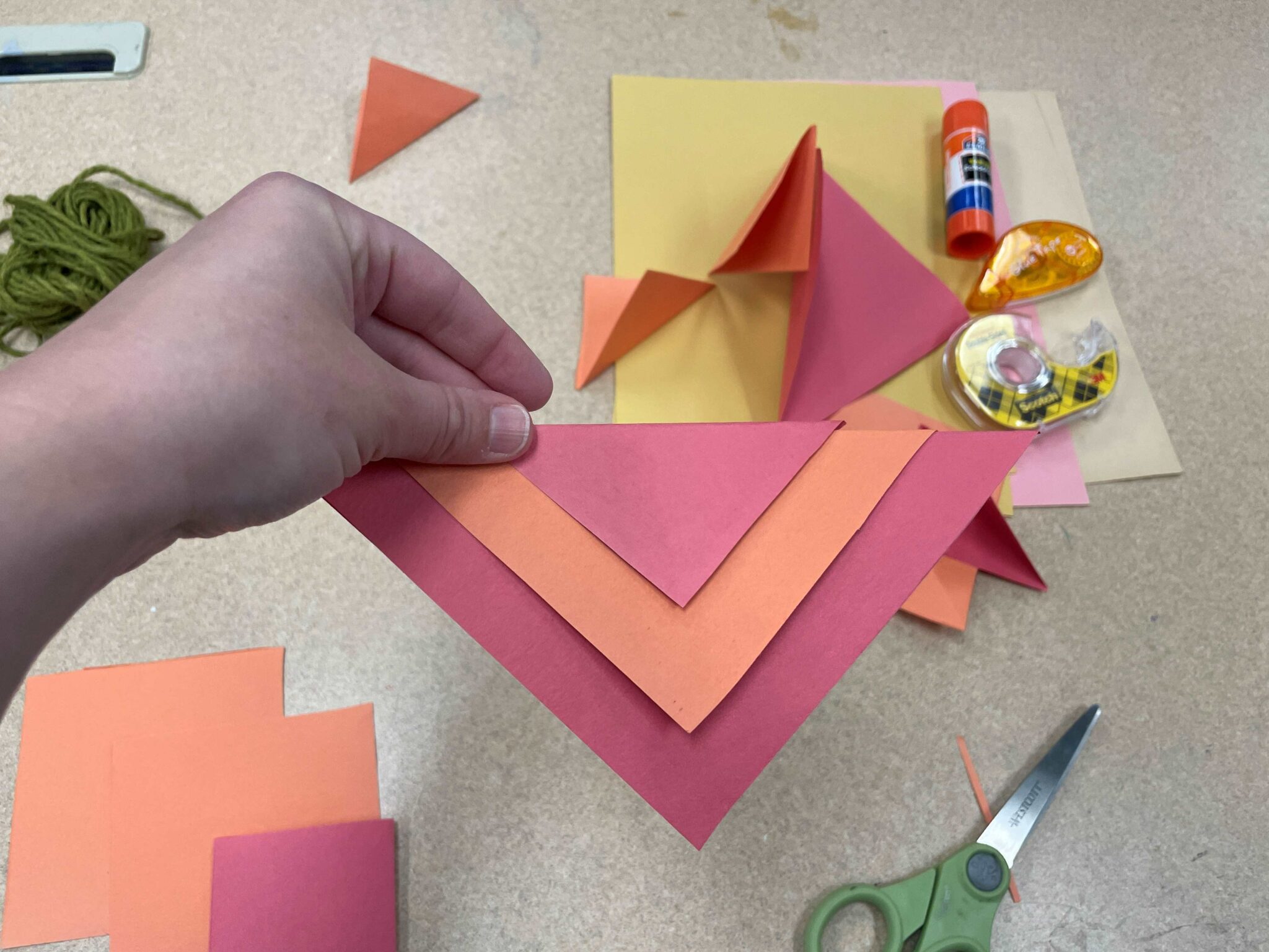 A white hand holds up three paper triangles layered on top of one another to create a red-orange-red colored flag. The triangles are arranged in a downward-pointing chevron pattern. In the background are two orange and one red squares of paper, which, when folded in half, could create the same triangle flag shape. There are other orange and red squares folded into triangles in the background as well, along with a ball of green twine, a pair of green scissors, and a variety of adhesives, including a glue stick, roll of double-sided tape, and a glue tape dispenser. 
