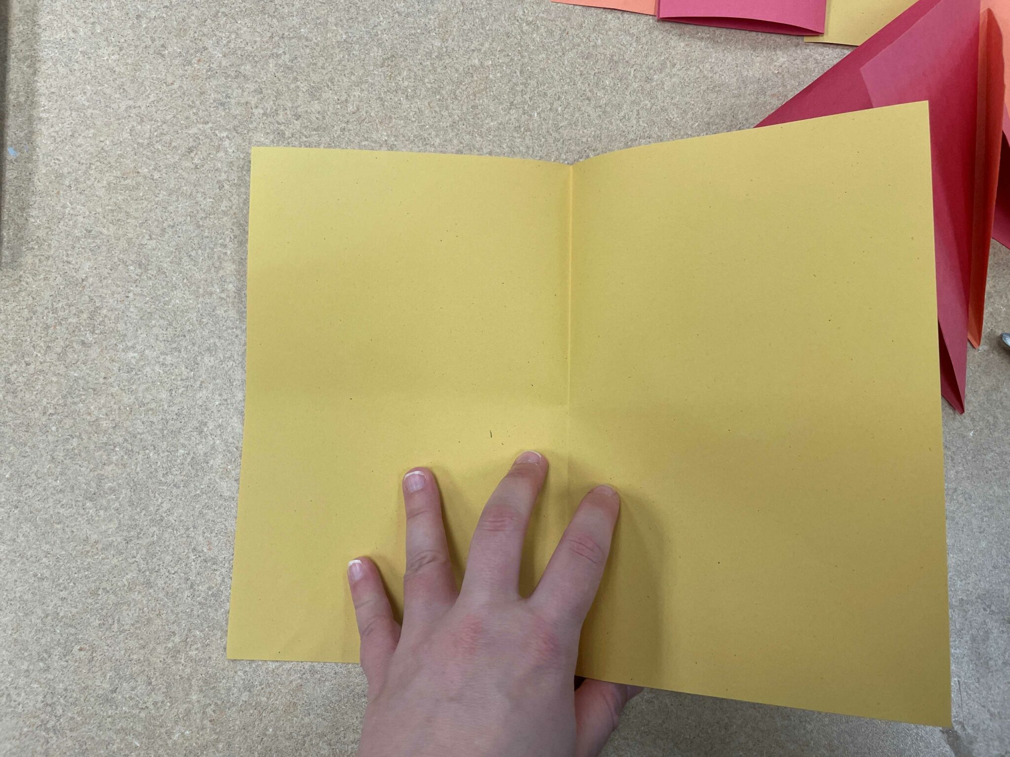 A piece of yellow paper with a crease down the middle, illustrating a "hamburger style" fold. A white hand holds the paper near the crease. 