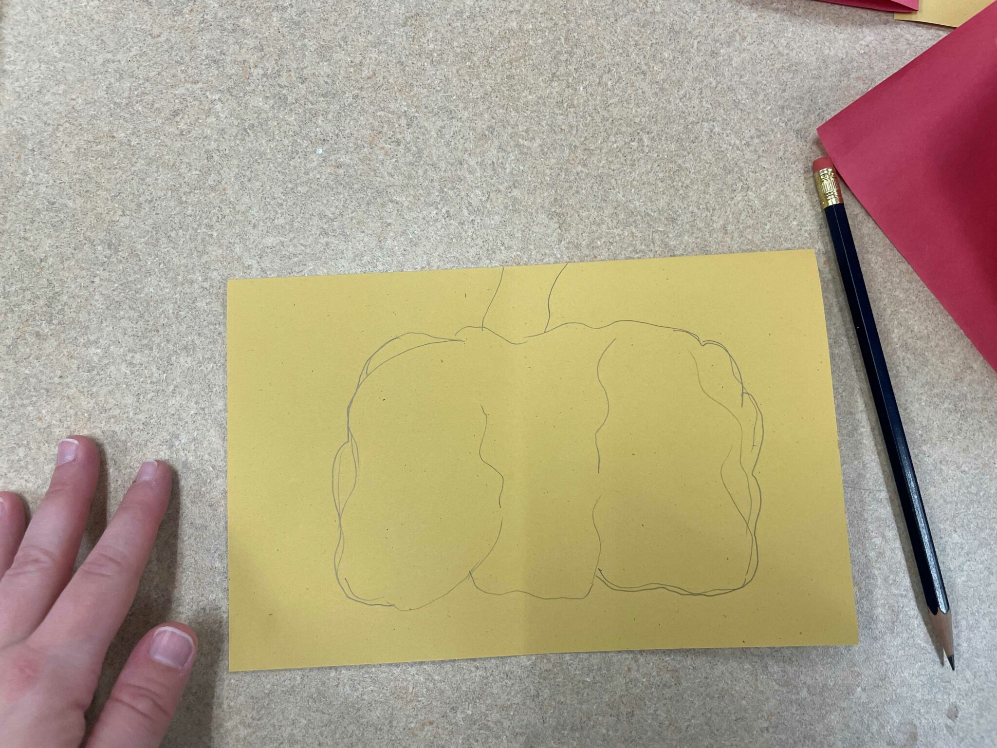 A half sheet of yellow paper next to a white hand. A large pumpkin has been drawn in light pencil on the paper. 