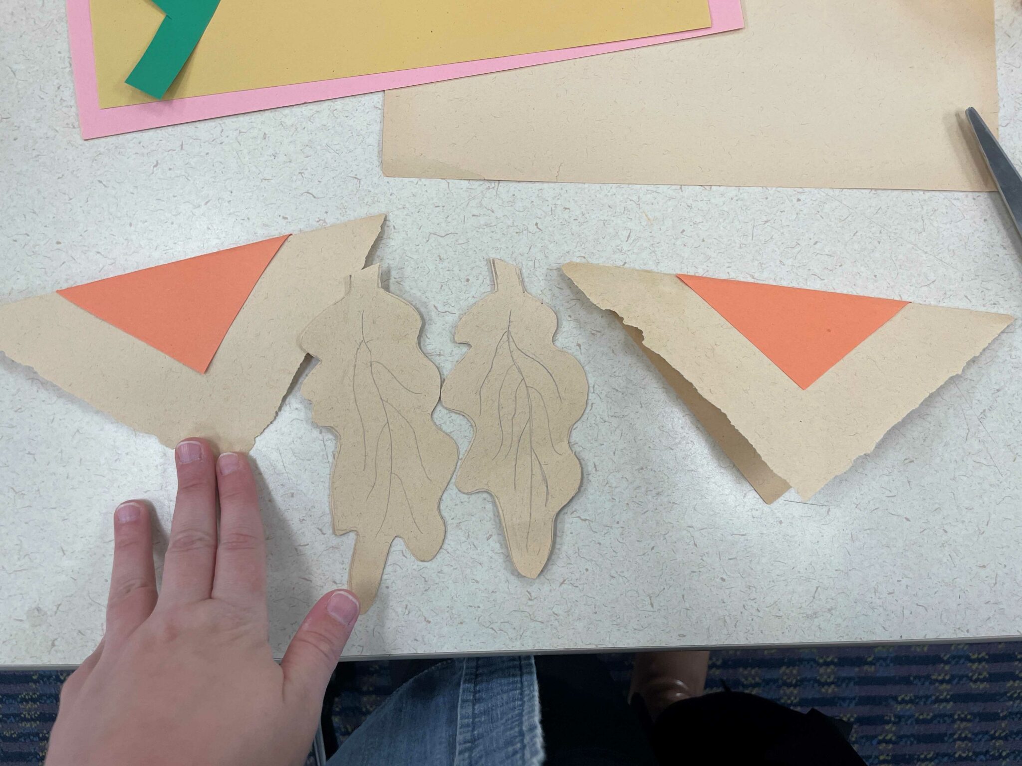 A white hand holds down a triangle paper flag made up of two colored triangles in brown and orange. The paper flag has a downward-pointing chevron pattern. Next to the flag are two oak leaves drawn onto brown paper and cutout. On the other side of the leaves is another brown and orange triangle flag. 