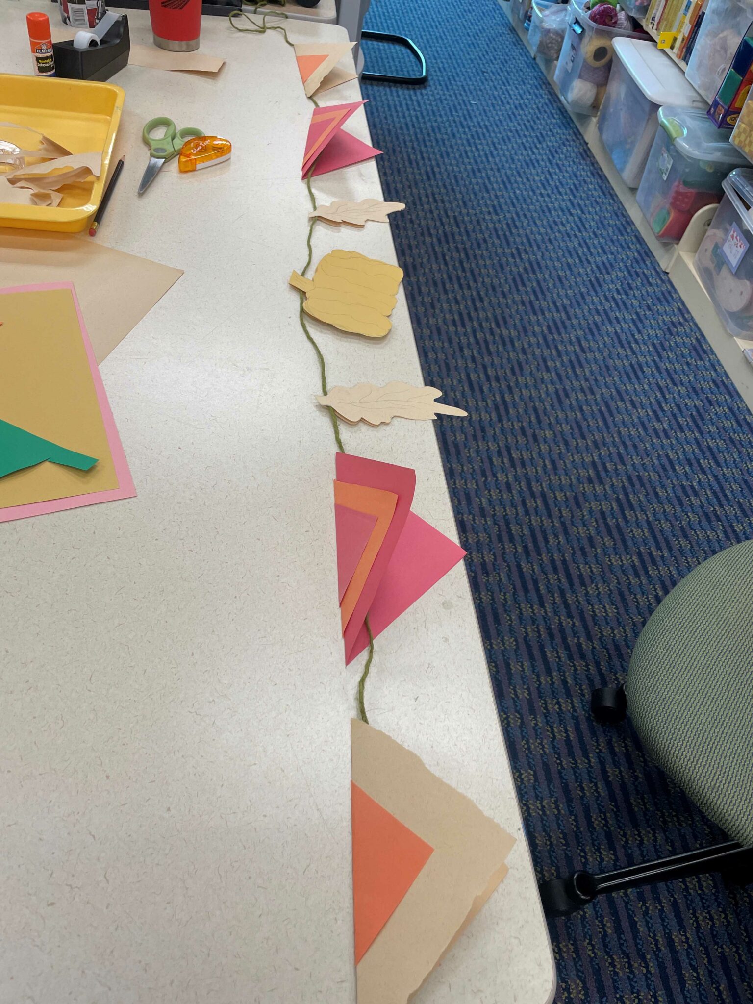 A couple feet of green twice unspooled with paper cutouts evenly spaced out along the twine. The cutouts include four triangle flags (two in brown and orange, and two in red-orange-red), a yellow pumpkin, and two brown oak leaves. 