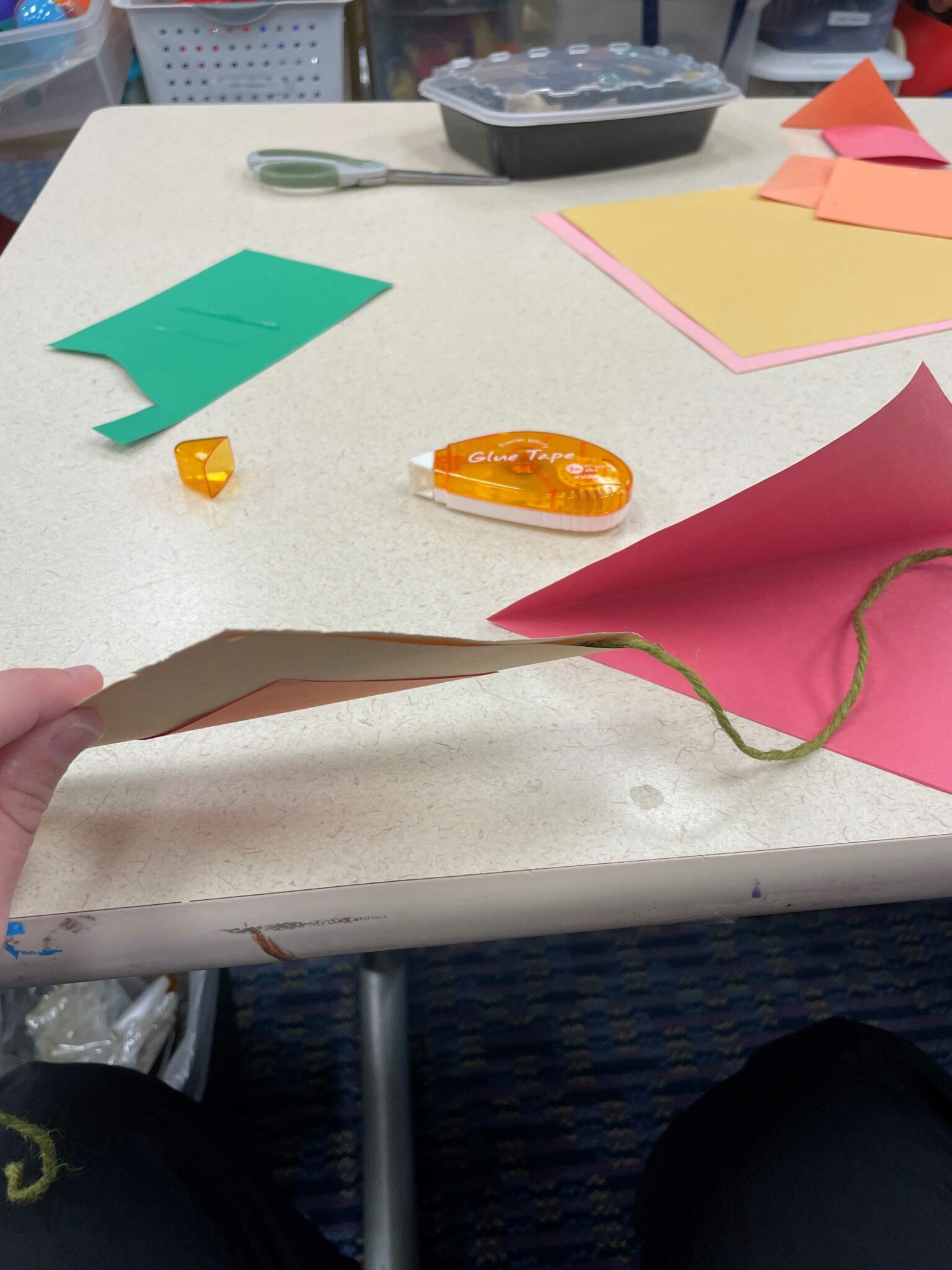 One of the brown and orange triangle flags held up by a white hand. The two sides of the paper triangle flag have been glued together around a long piece of green twine, to fasten the paper cutout to the beginning of the bunting. 