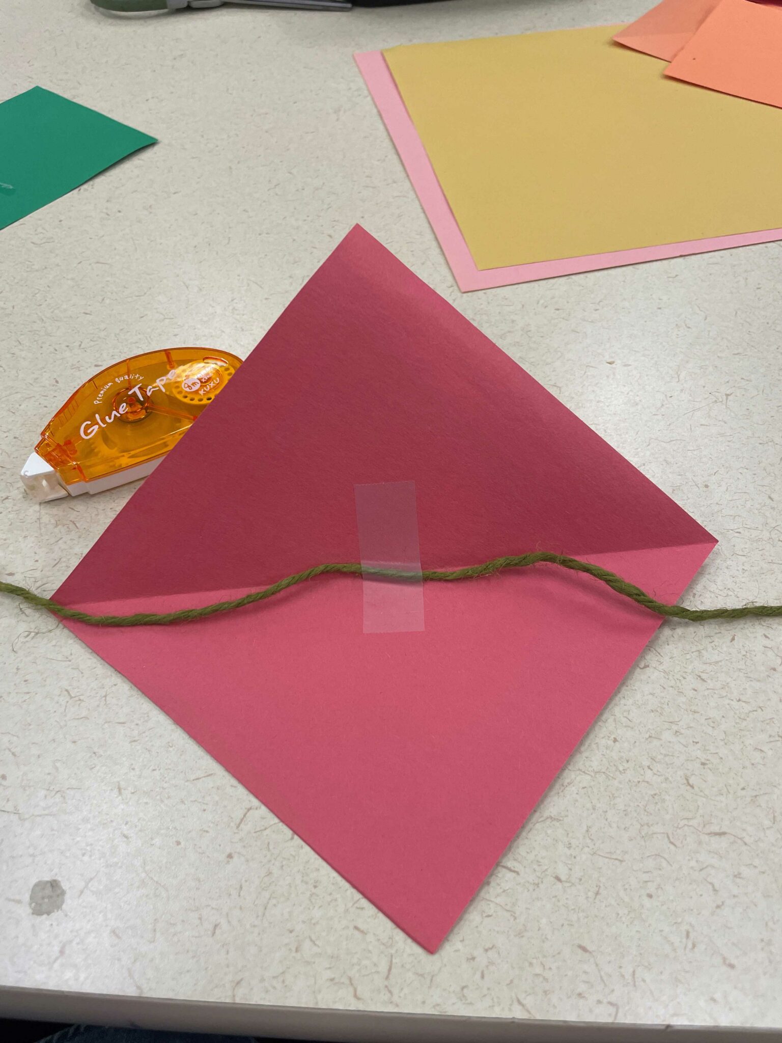 One of the red-orange-red triangle flags, opened up to reveal a red diamond shape with a horizontal crease running down the middle. A piece of green twine has been run under the center crease, and a piece of tape has been placed on this twine to fasten the flag to it. 
