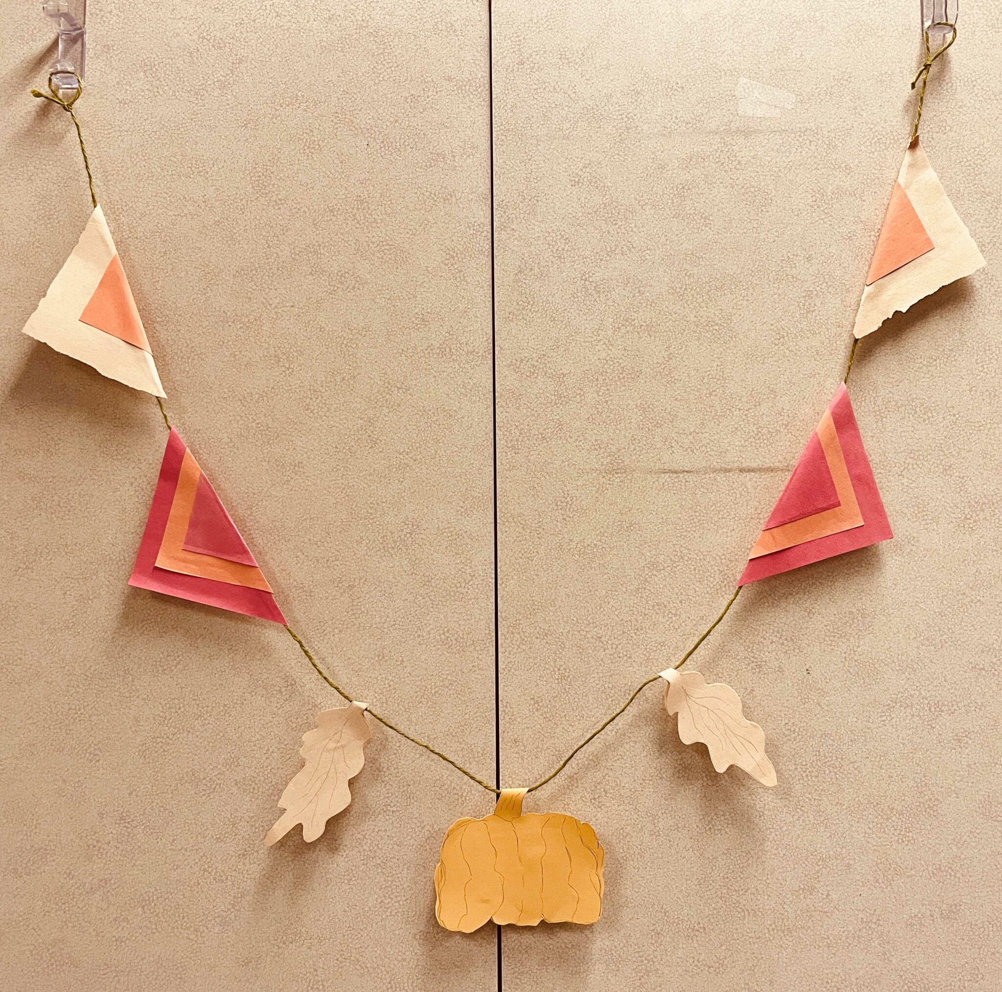 Image of a handmade bunting with paper cutouts taped to green twine. The paper cutouts include a two brown and orange triangle flags, two red-orange-red triangle flags, two brown oak leaves, and a yellow-orange pumpkin. The bunting hangs from two clear hooks over two cabinet doors.