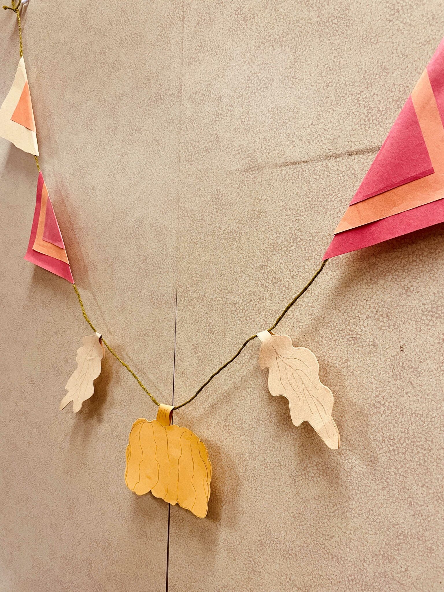 Image of a handmade bunting with paper cutouts taped to green twine. The paper cutouts include a brown and orange triangle flag, two red-orange-red triangle flags, two brown oak leaves, and a yellow-orange pumpkin. 