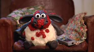 A close-up of Timmy, a Shaun the Sheep claymation character, sitting on a reddish brown armchair with a floral blanket behind him. Timmy is a black lamb with white wool and has red pizza sauce smeared all over his head and torso. 