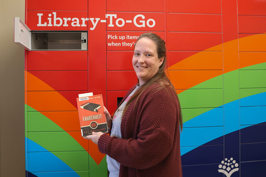 Smiling person holding a book by a Library-To-Go locker